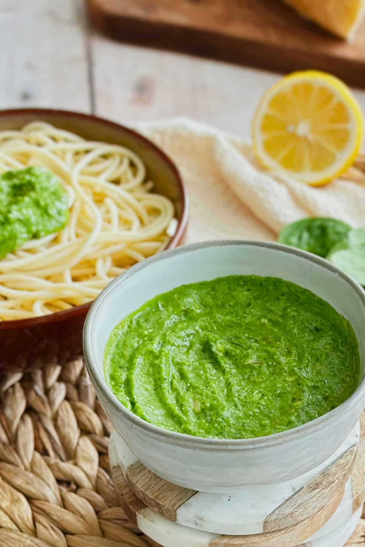A bowl of vibrant green roasted garlic and spinach pesto sits beside a plate of spaghetti with a lemon wedge and spinach leaves in the background.