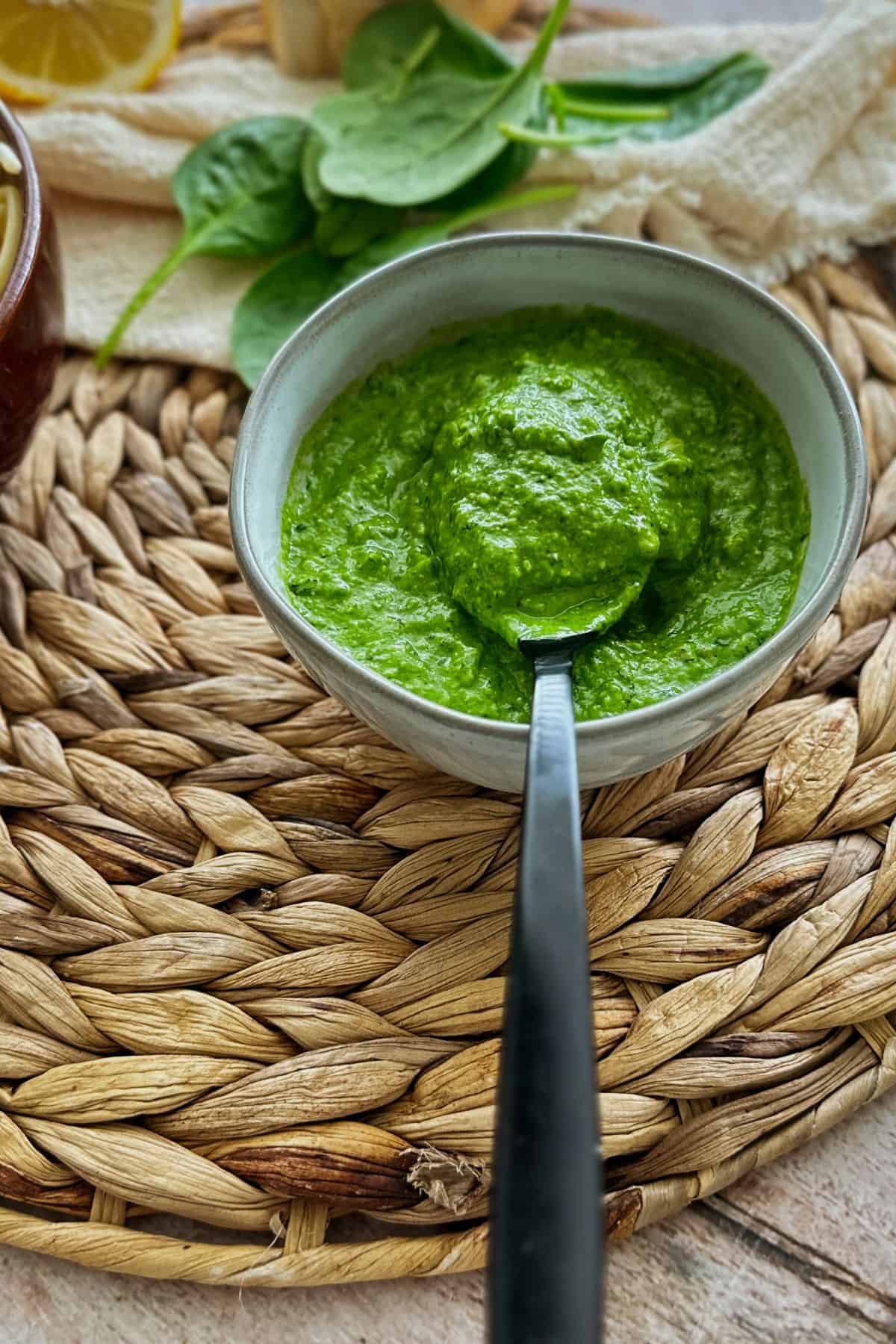 A bowl of bright green roasted garlic and spinach pesto with a spoon, next to a dish of spaghetti and fresh spinach leaves on a woven placemat.