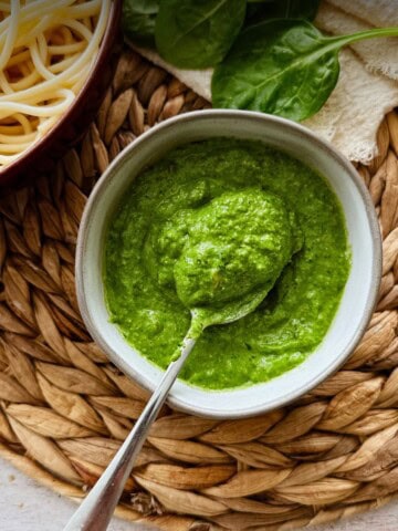 A bowl of vibrant green roasted garlic and spinach pesto with a spoon, beside a bowl of spaghetti and fresh spinach leaves on a light napkin.