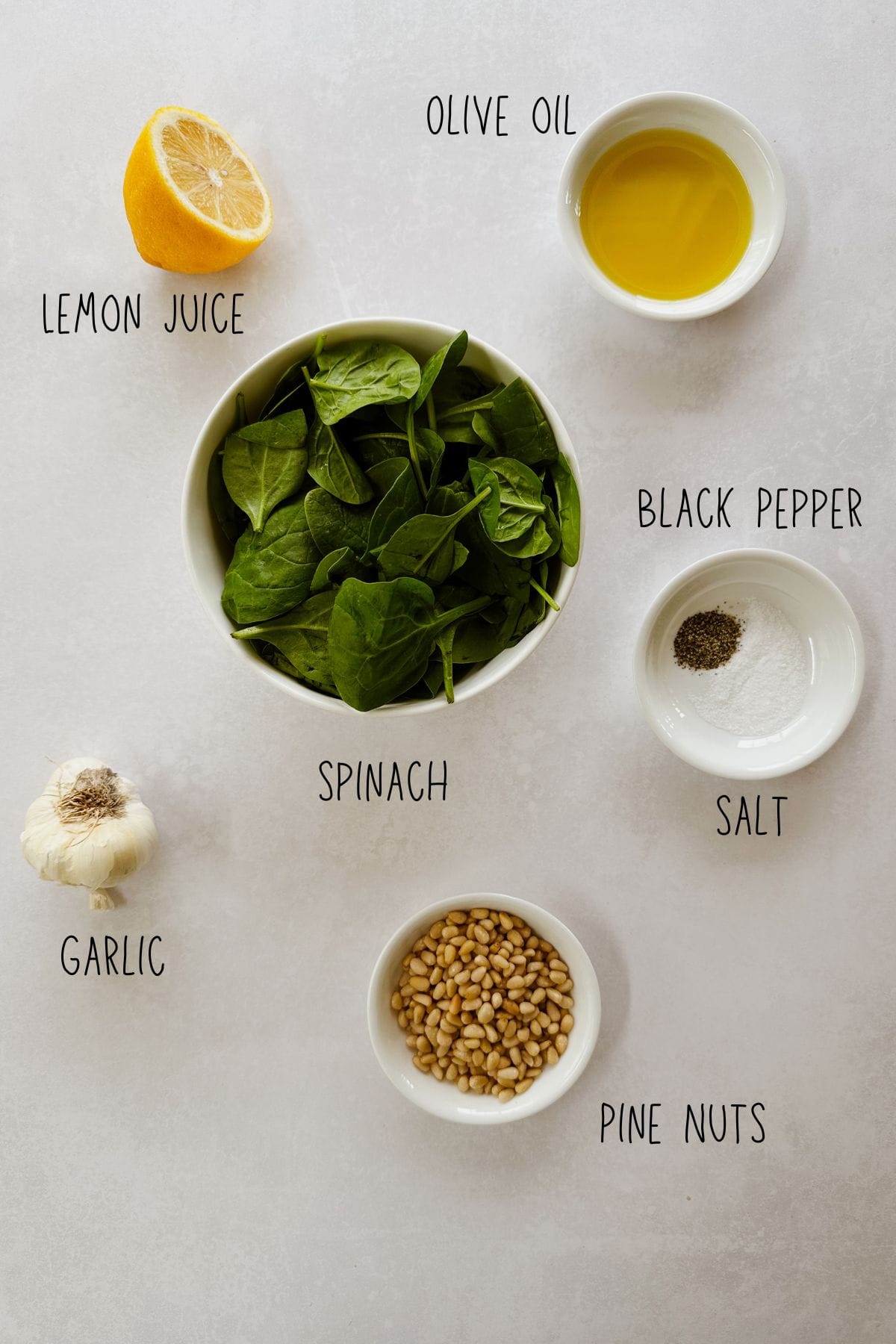 An overhead view of ingredients for roasted garlic and spinach pesto arranged on a light surface, including fresh spinach, olive oil, lemon juice, garlic, pine nuts, salt, and black pepper. Each ingredient is labeled with text.
