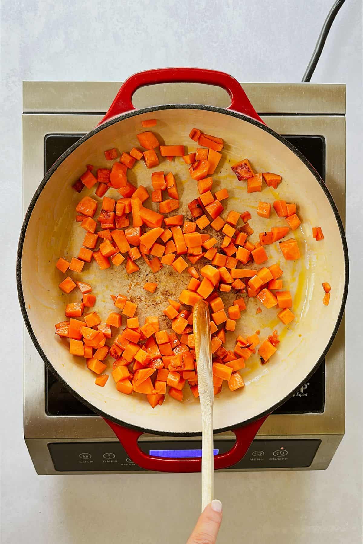 A red Dutch oven sits on a stovetop, filled with cubed orange sweet potatoes sizzling in olive oil. A wooden spoon stirs them as they start to brown slightly around the edges.
