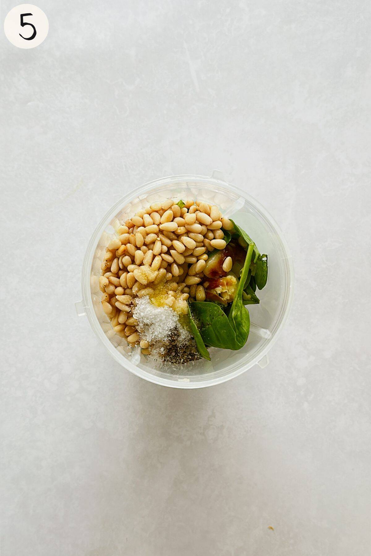 Overhead view of a blender cup filled with pesto ingredients - pine nuts, spinach, roasted garlic, salt, pepper, and lemon juice ready to blend.
