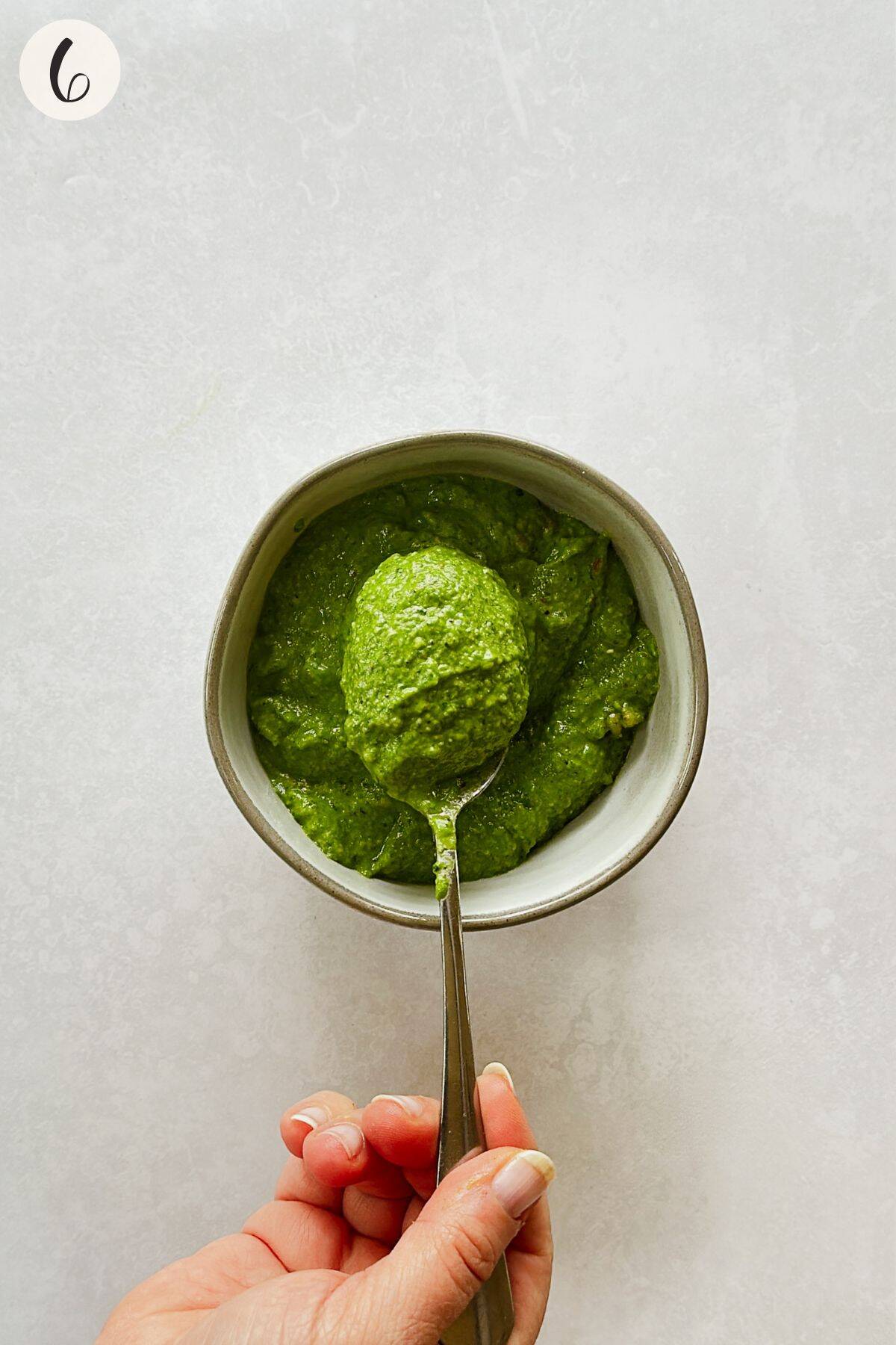A hand holds a spoonful of bright green spinach pesto over a bowl, showing its creamy texture and vibrant color after blending.