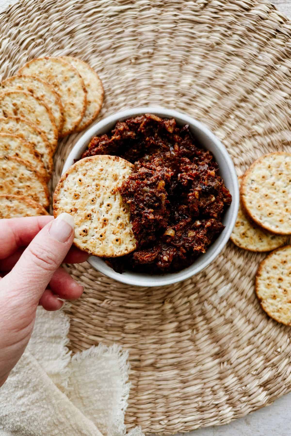 A hand dipping a cracker into a bowl of chunky sun-dried tomato spread, surrounded by more round crackers on a woven placemat.