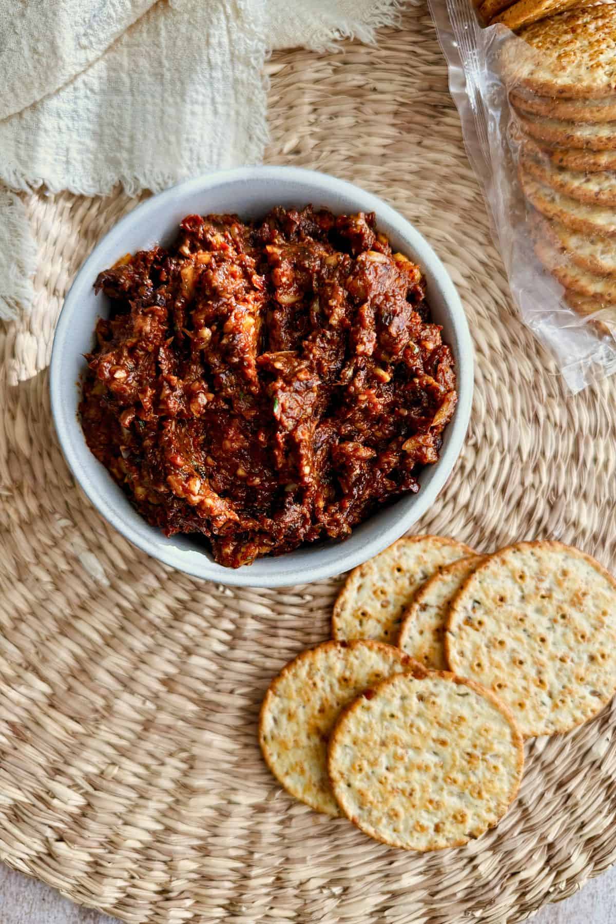 A bowl full of sun dried tomato spread surrounded by round crackers.