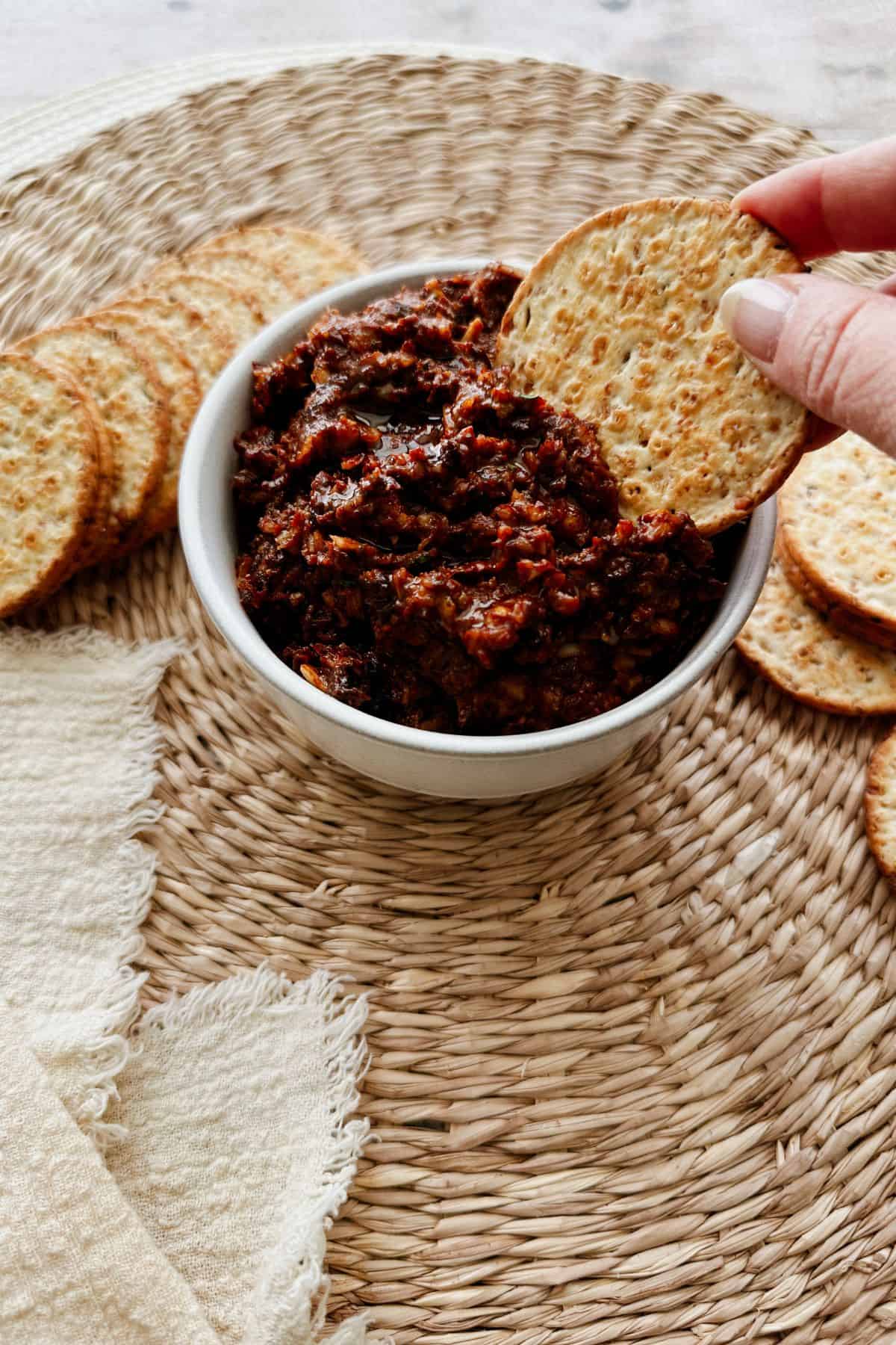 An angled view of a hand dipping a cracker into a bowl of chunky sun-dried tomato spread, surrounded by more round crackers on a woven placemat.