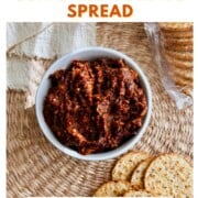 A bowl of chunky sun-dried tomato spread surrounded by round crackers on a woven placemat. Text on the image reads “How to Make Sun-Dried Tomato Spread – Great for an appetizer board!”