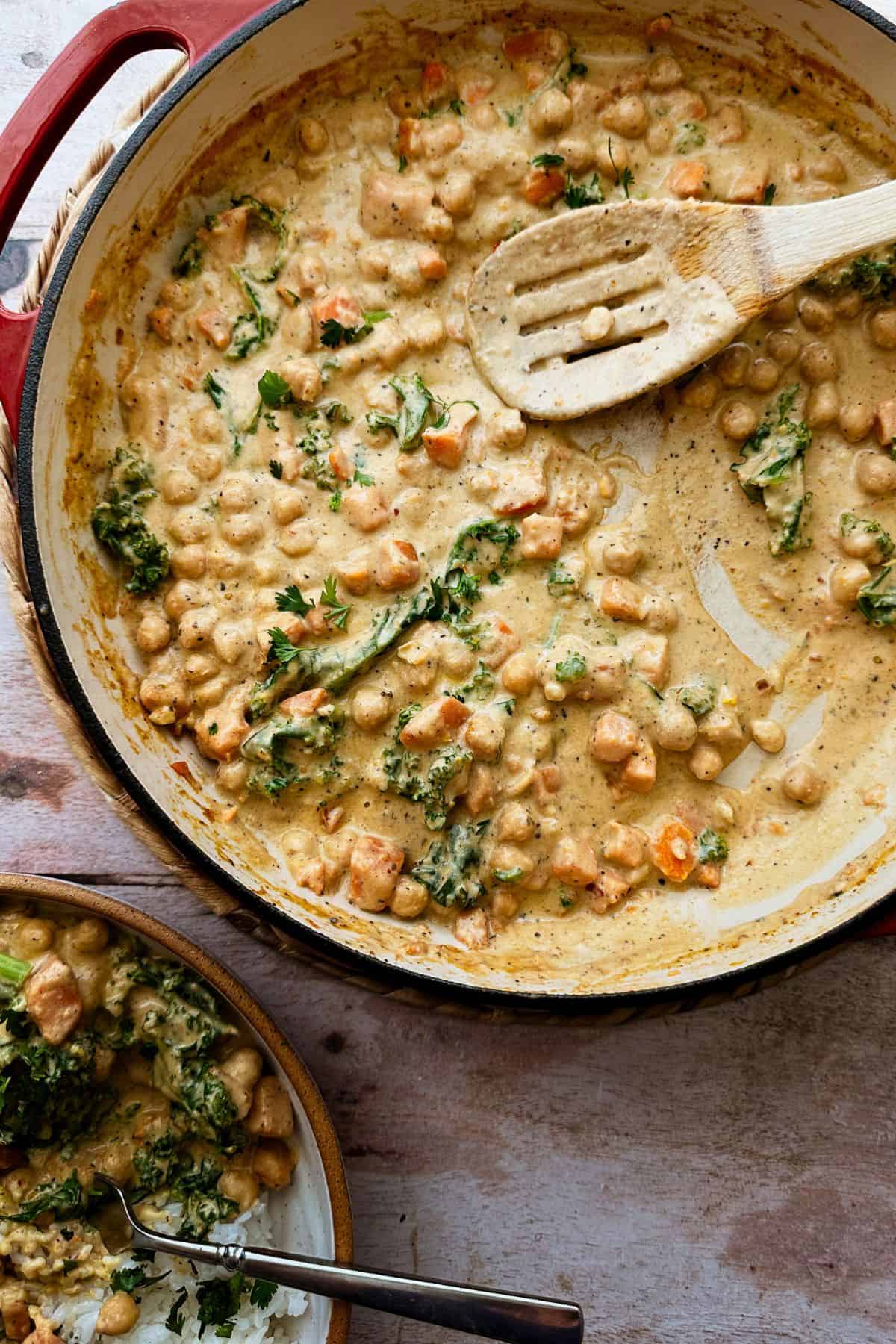 A close-up of creamy chickpea and sweet potatoes in the red pan. Wooden serving spoon is layed in the pan.