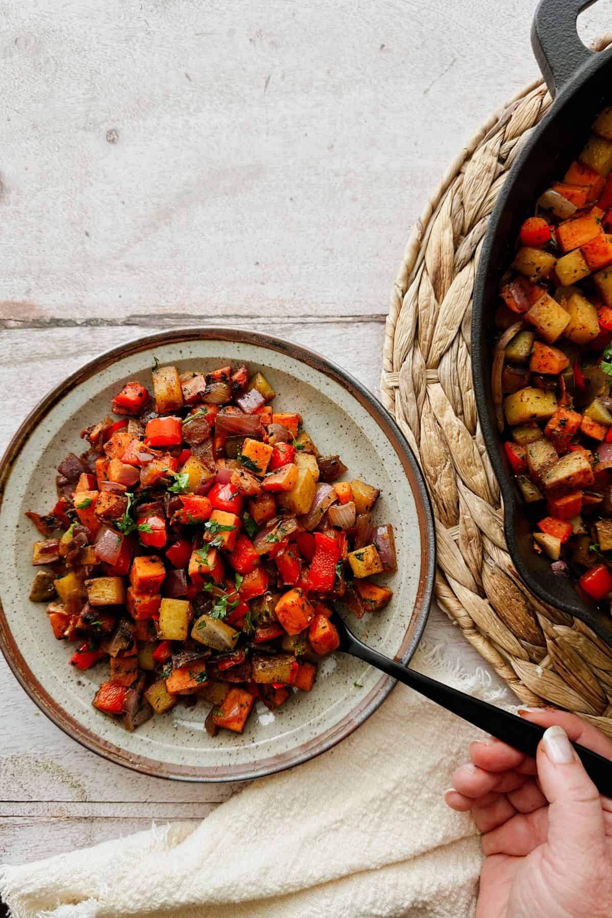 A plate of colorful sweet potato and russet hash with red peppers, onions, and herbs. A hand holds a black spoon beside a cast-iron skillet of the same dish.