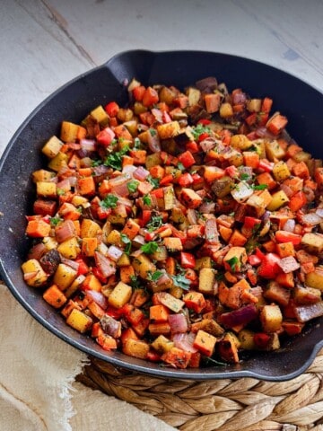 A skillet filled with sweet potato and russet hash with red peppers, onions, and herbs. The dish rests on a woven mat with a cream-colored cloth.