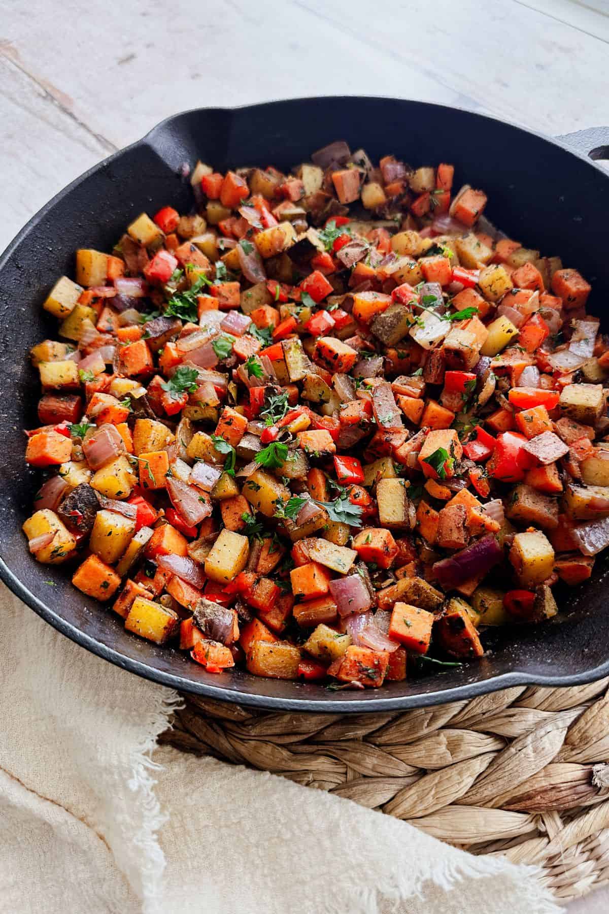 A skillet filled with sweet potato and russet hash with red peppers, onions, and herbs. The dish rests on a woven mat with a cream-colored cloth.