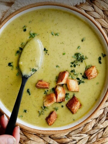 Bowl of creamy zucchini soup topped with fresh thyme and golden croutons, served with a spoon on a woven placemat.