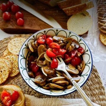 Bowl of mushroom salad with onions and cherry tomatoes in a tangy dressing, surrounded by crackers, sliced bread, and fresh tomatoes on a cutting board.