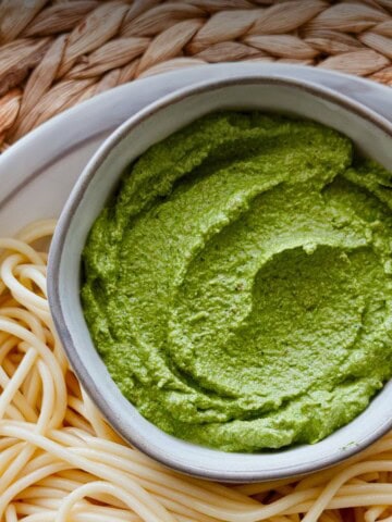 Overhead view of a bowl filled with creamy walnut and kale pesto next to a plate of plain spaghetti, ready to be mixed and served.