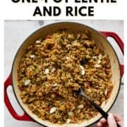 A large red Dutch oven filled with flavorful one-pot lentils and rice, garnished with herbs and sliced almonds, with a hand holding a black spoon ready to serve.