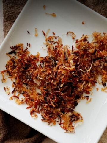 Golden, crispy fried shallots cooling on a white plate, ready to use as a crunchy topping or garnish.