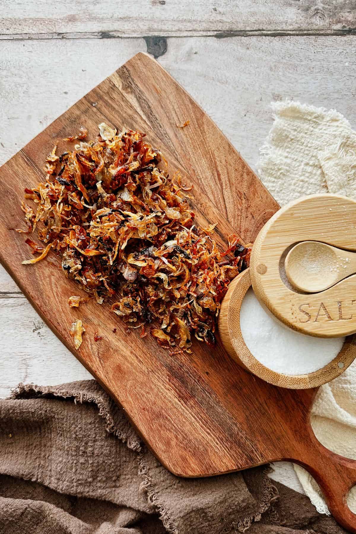 Golden crispy shallots on a wooden board beside a small salt container, ready to season or use as a topping.
