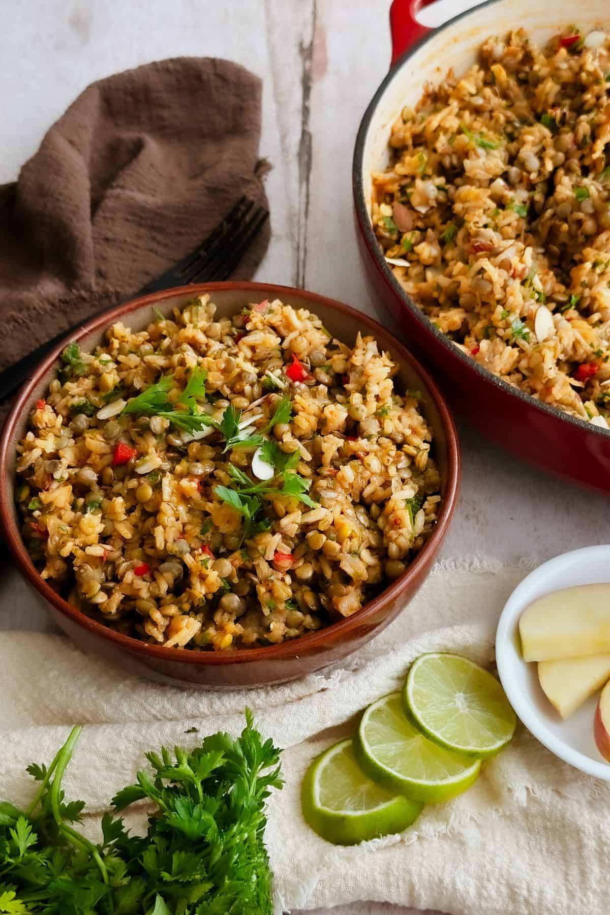 A close-up of one-pot lentils and rice served in a brown bowl, garnished with fresh parsley. The red Dutch oven with more of the dish is in the background, alongside lime slices, herbs, and a beige napkin on a rustic tabletop.
