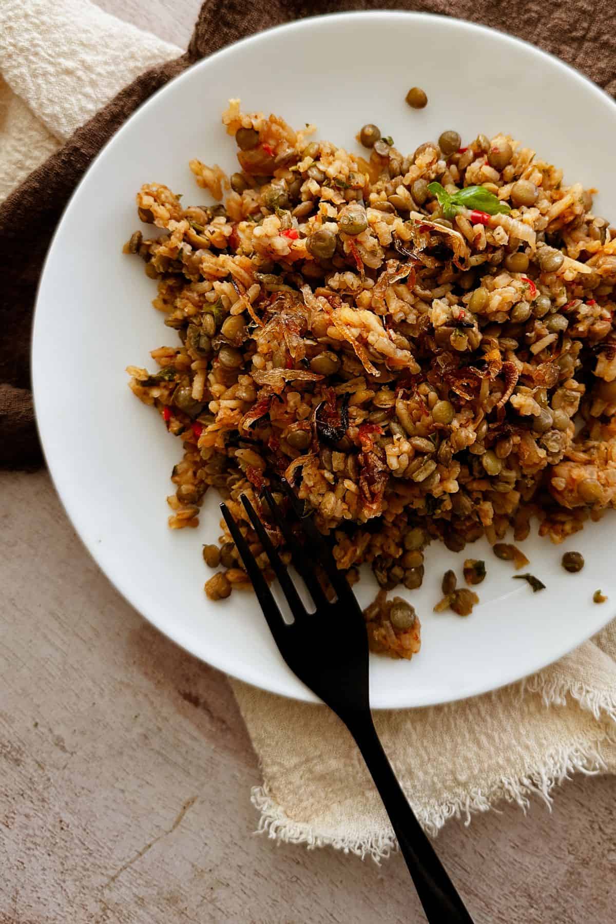  A white plate filled with one-pot lentils and rice, garnished with crispy shallots and fresh herbs, placed on a beige cloth with a brown napkin and black fork beside it.