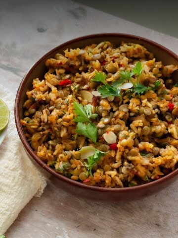 A bowl of one-pot lentils and rice garnished with fresh parsley and red pepper pieces, placed on a rustic table with lime slices and a cream-colored napkin beside it.