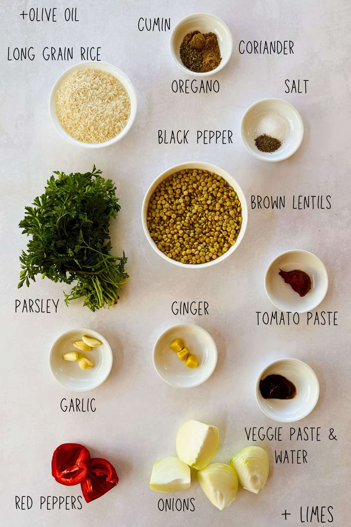 Ingredients for one-pot lentils and rice, including brown lentils, long-grain rice, onions, red peppers, garlic, ginger, tomato paste, vegetable paste, parsley, lime, cumin, coriander, oregano, salt, and black pepper arranged on a light surface.