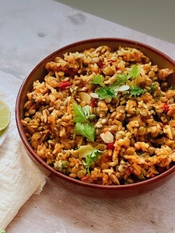 A bowl of one-pot lentils and rice garnished with fresh parsley and red pepper pieces, placed on a rustic table with lime slices and a cream-colored napkin beside it.