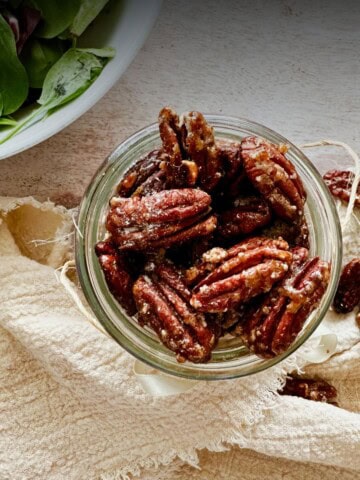 Overhead view of a jar filled with candied cinnamon pecans on a linen cloth, with a few pecans scattered nearby.