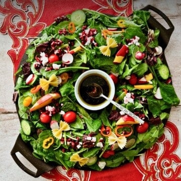 Overhead view of a Christmas wreath salad on a black round platter, mixed greens arranged in a ring with colorful toppings and a bowl of dark dressing in the center.