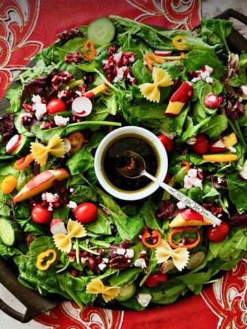 Overhead view of a Christmas wreath salad on a black round platter, mixed greens arranged in a ring with colorful toppings and a bowl of dark dressing in the center.