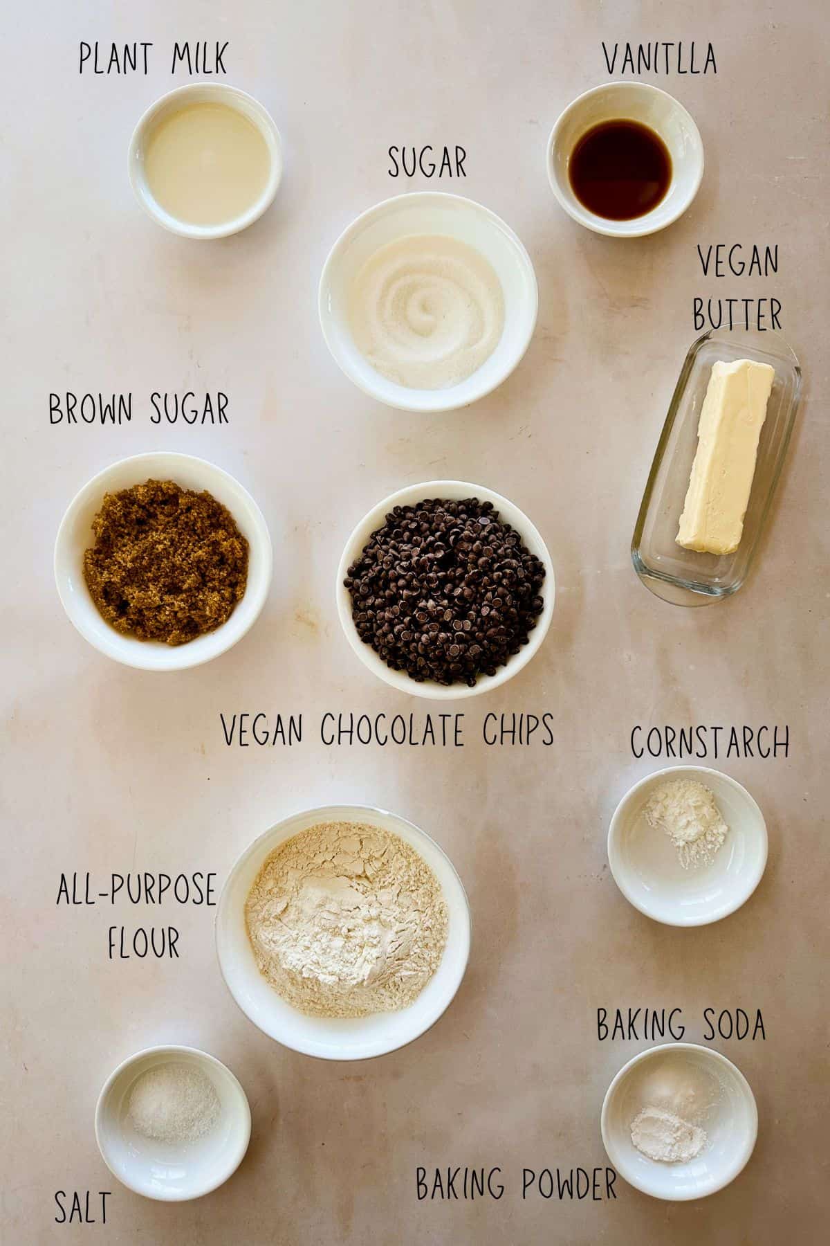 Flat lay of vegan cookie ingredients: plant milk, vanilla, sugar, brown sugar, vegan butter, chocolate chips, flour, cornstarch, baking soda, salt.