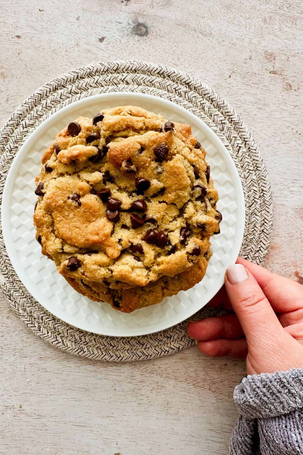 Large vegan chocolate chip cookie on a white plate, held by a hand.