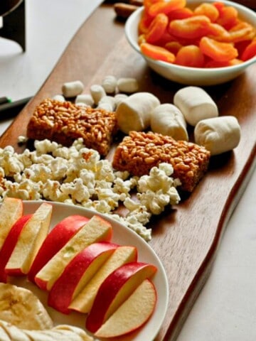 Wooden snack board with apple slices, popcorn, marshmallows, rice crispy treats, and a bowl of dried apricots.