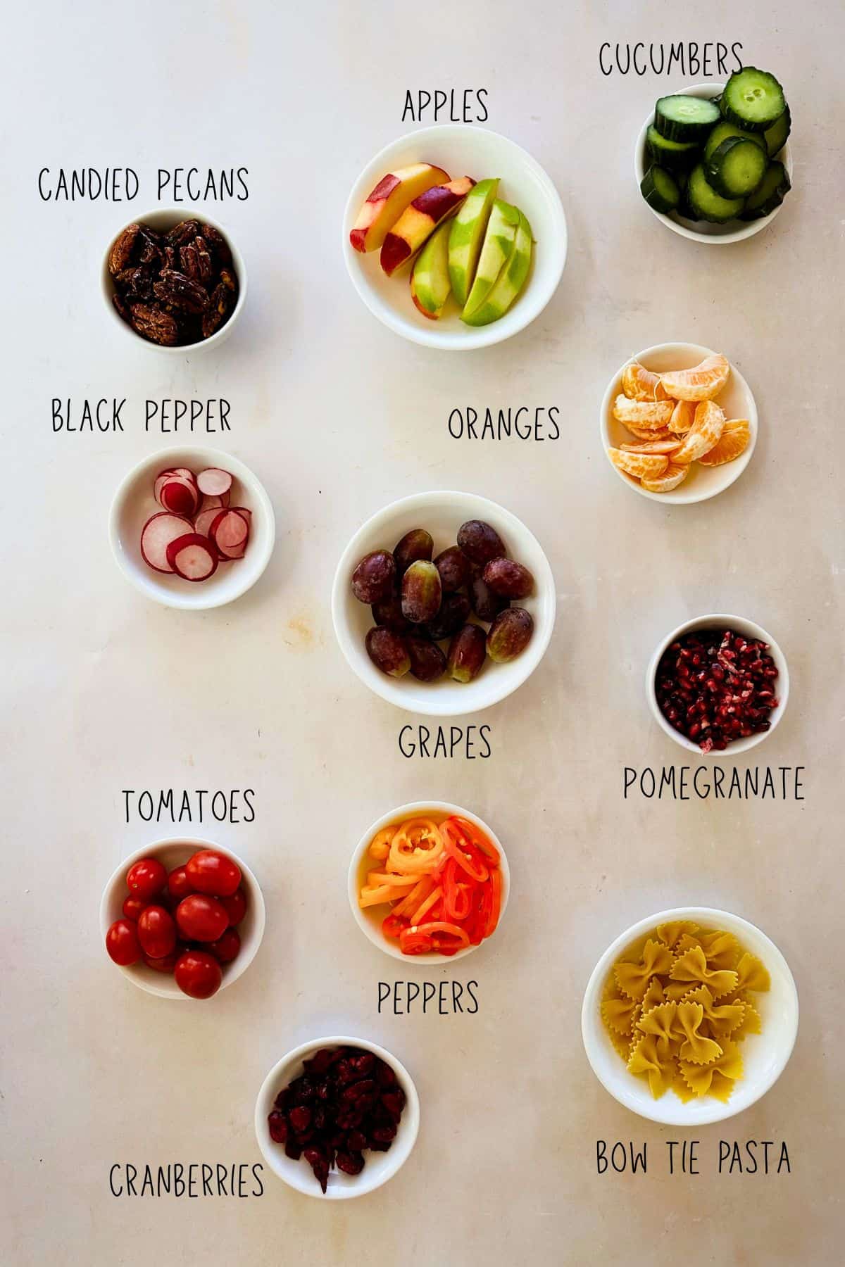 Overhead photo of small white bowls of salad ingredients on a light surface, labeled candied pecans, apples, cucumbers, black pepper (sliced radishes), oranges, grapes, pomegranate arils, tomatoes, peppers, cranberries, and bow tie pasta.