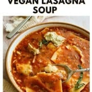 Overhead view of a bowl of vegan lasagna soup with broken lasagna noodles in a tomato-lentil broth, topped with vegan ricotta and fresh herbs, with a spoon nearby.