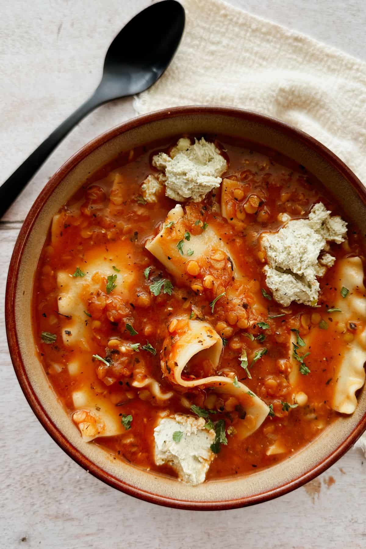 Overhead view of a bowl of vegan lasagna soup with broken lasagna noodles in a tomato-lentil broth, topped with vegan ricotta and fresh herbs, with a spoon nearby.