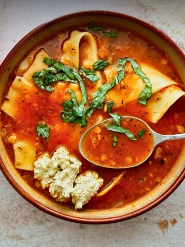 Overhead view of a bowl of vegan lasagna soup with wide lasagna noodles in a tomato-lentil broth, topped with fresh basil and vegan ricotta, with a spoon in the bowl.