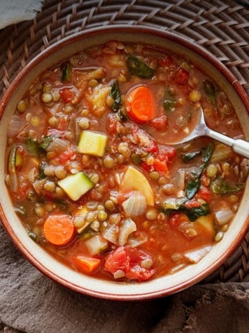 Overhead view of a bowl of lentil vegetable soup with tomatoes, carrots, zucchini, onions, and greens in a red broth, with a spoon in the soup.