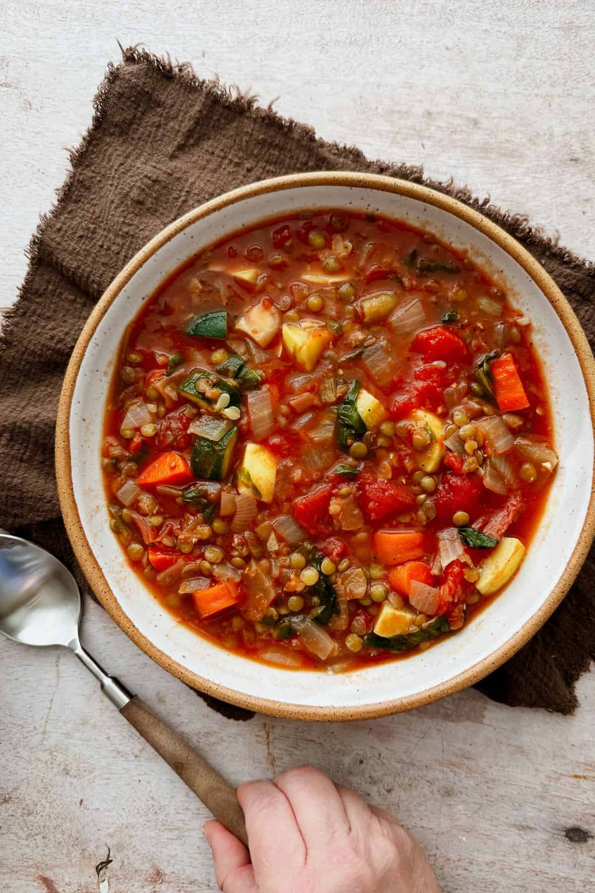 Top-down view of a bowl of lentil vegetable soup with tomatoes, carrots, zucchini, onions, and greens, with a spoon beside the bowl.