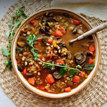A close-up of hearty mushroom barley soup in a rustic bowl, topped with sprig of fresh oregano with a napkin on the side.