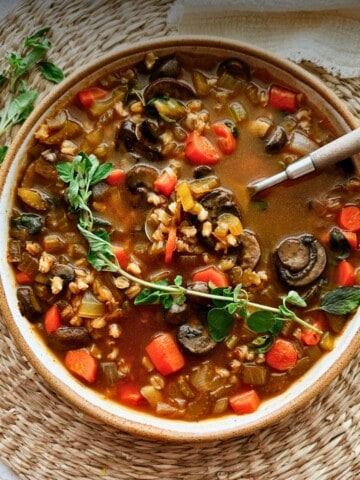 A close-up of hearty mushroom barley soup in a rustic bowl, topped with sprig of fresh oregano with a napkin on the side.