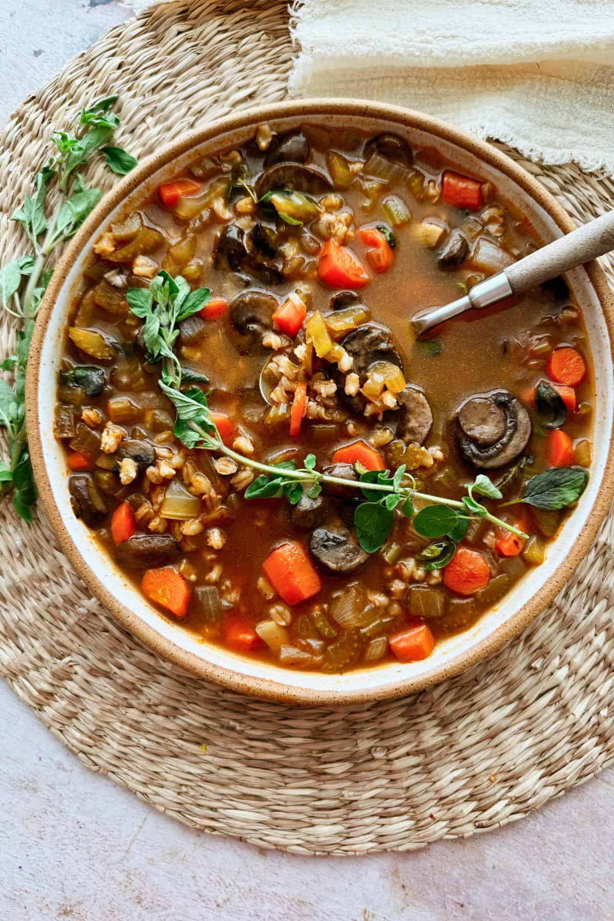 A close-up of hearty mushroom barley soup in a rustic bowl, topped with sprig of fresh oregano with a napkin on the side.