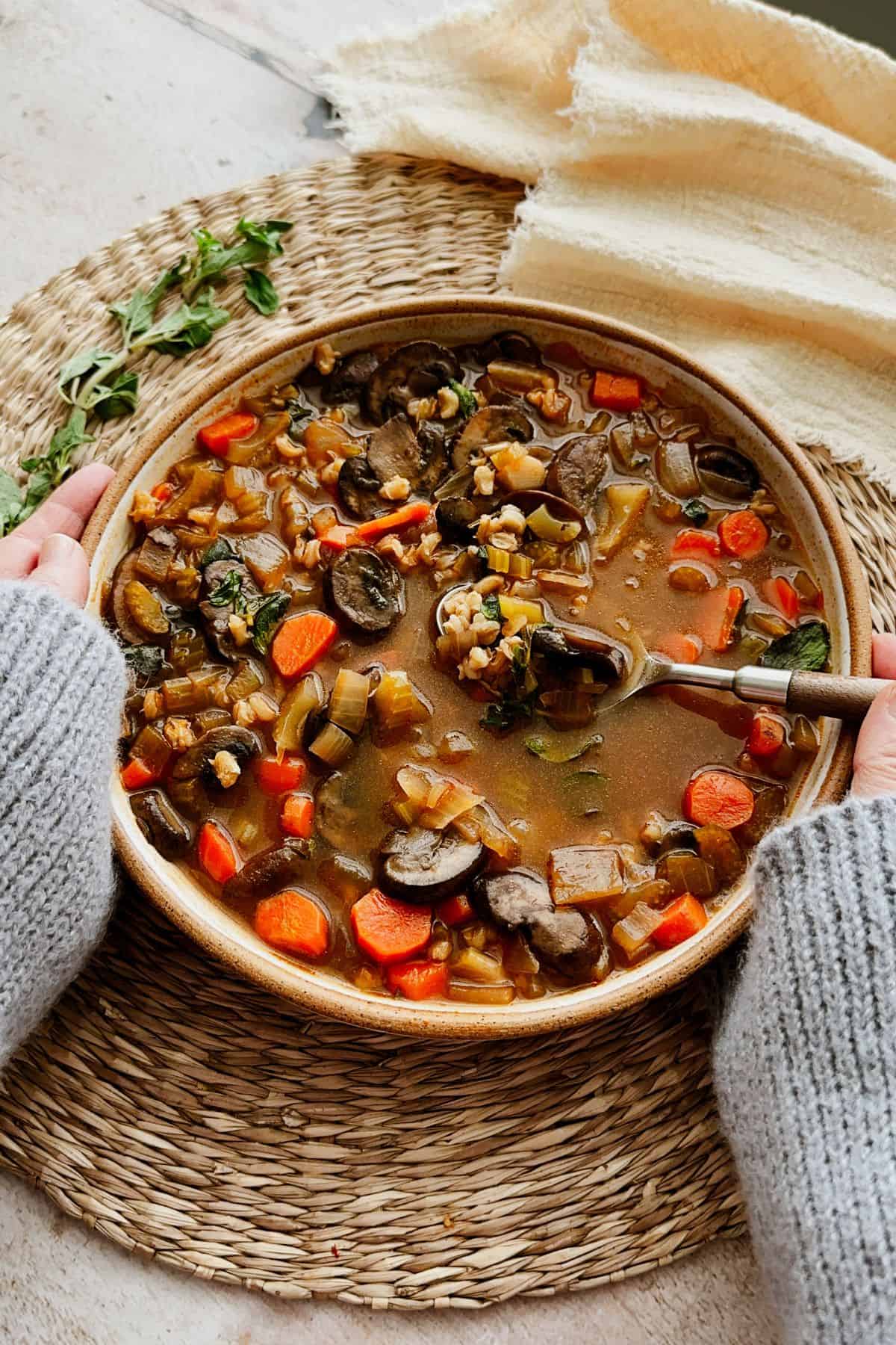 A cozy bowl of mushroom barley soup topped with fresh oregano. A hand holds the bowl while a spoon scoops up a bite.
