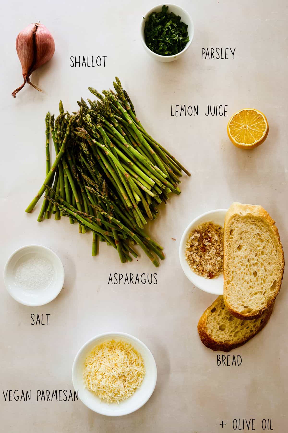 Flat lay of ingredients for vegan asparagus gratin including fresh asparagus, a shallot, chopped parsley, lemon juice, olive oil, salt, sourdough bread slices, homemade breadcrumbs, and grated vegan parmesan.