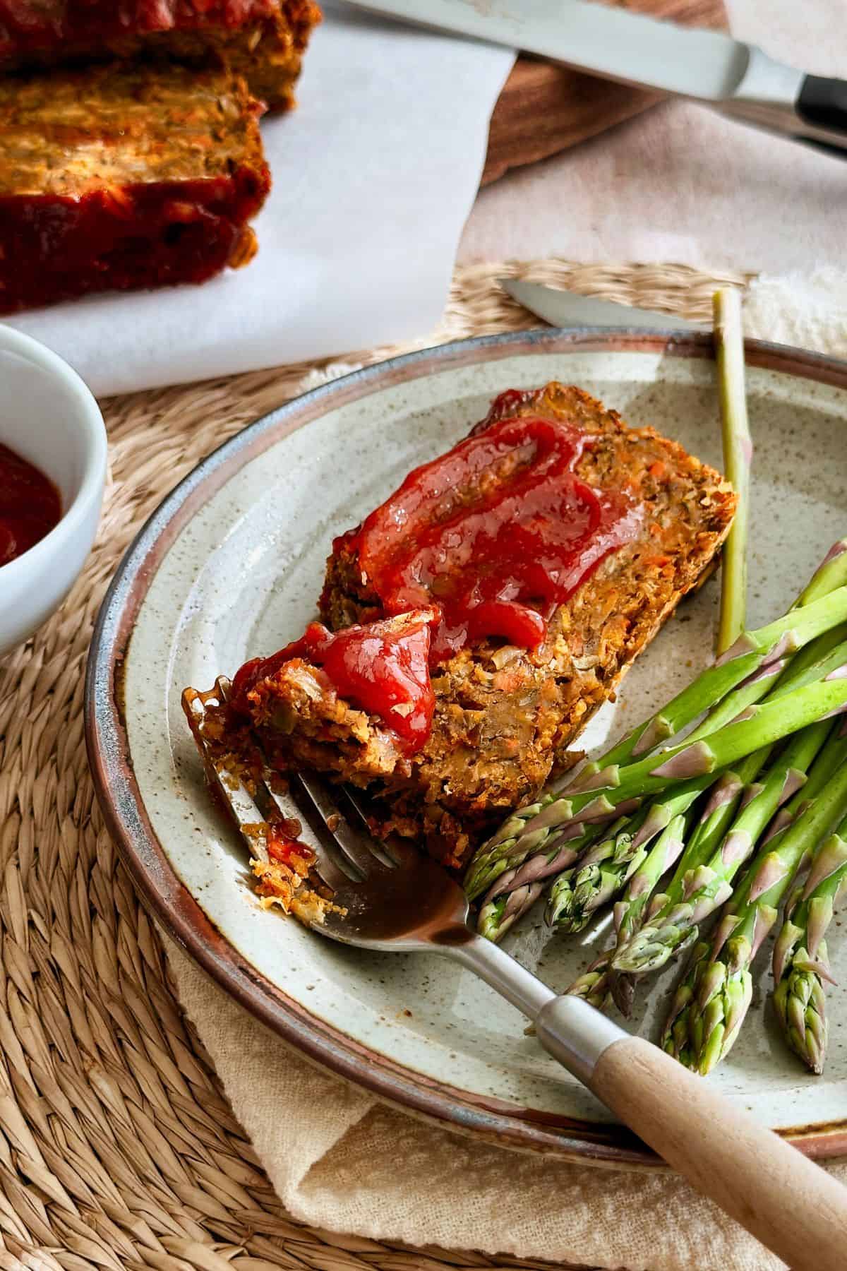 A plated slice of vegan lentil loaf with ketchup glaze served alongside roasted asparagus on a ceramic plate, ready to eat.