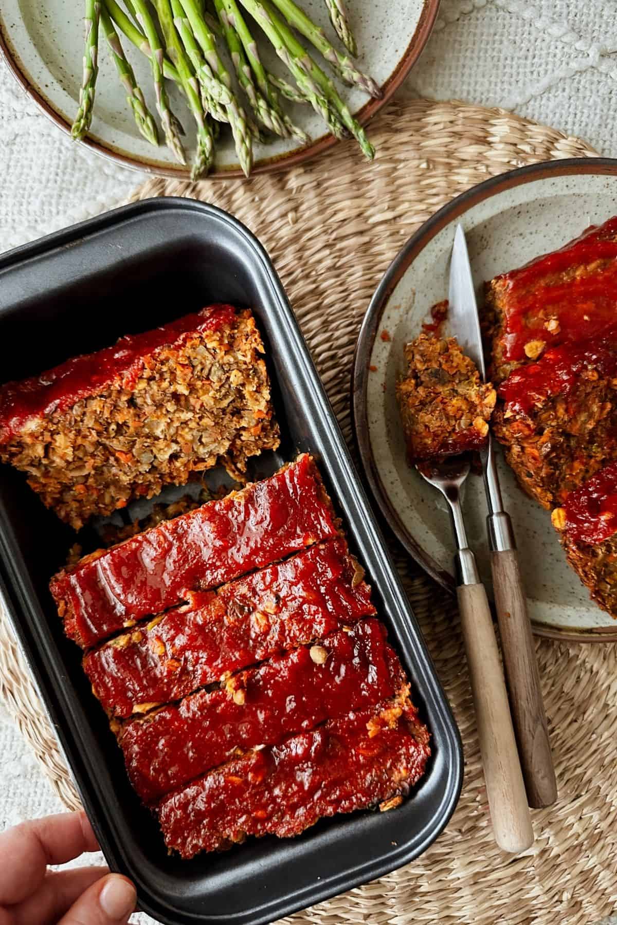 Sliced vegan lentil loaf in a black loaf pan, topped with a glossy ketchup glaze, with plated slices and roasted asparagus arranged on a woven placemat nearby.