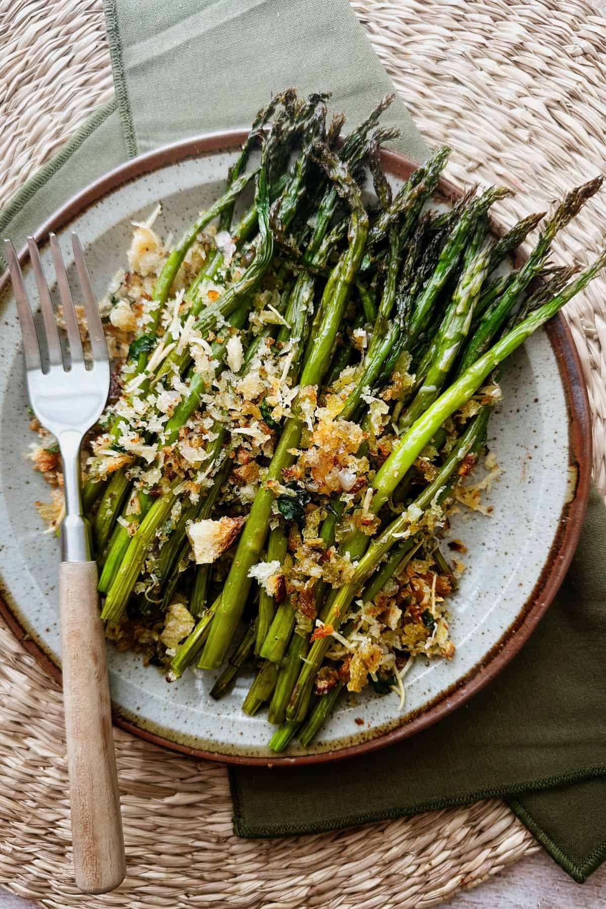 A plate of vegan asparagus gratin with roasted asparagus spears topped with crisp breadcrumbs and vegan parmesan, served on a ceramic plate with a fork resting alongside.