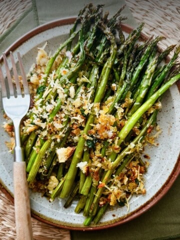 A plate of vegan asparagus gratin with tender roasted asparagus spears topped with crispy golden breadcrumbs, vegan parmesan, and herbs, served with a fork on a rustic ceramic plate.