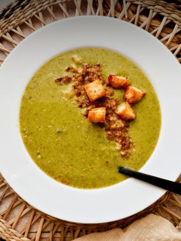 Bowl of creamy green broccoli soup on a woven placemat, topped with crispy croutons and breadcrumbs, with a spoon resting in the bowl.
