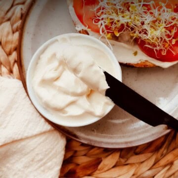 Small bowl of creamy vegan tofu mayo with a knife spread beside an open-faced sandwich with the mayo and topped with tomato and sprouts.
