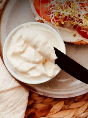 Small bowl of creamy vegan tofu mayo with a knife spread beside an open-faced sandwich with the mayo and topped with tomato and sprouts.